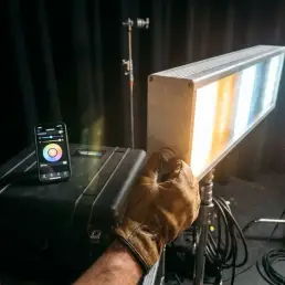 Cinematographer adjusting a bi-colour LED panel on a dark film set, warm amber and cool daylight tones glowing from the aluminum fixture.