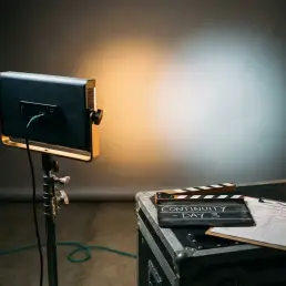 Bi-colour LED panel casting warm amber and cool daylight on a grey studio wall, with clapperboard and production schedule on a nearby equipment case.