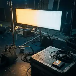 Bi-colour LED panel transitioning from warm amber to cool daylight white on a C-stand at a dimly lit film set, voltage meter and cables nearby.