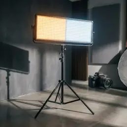 Bi-colour LED panel light showing warm amber and cool daylight tones on a studio stand, with camera and reflector visible in background.
