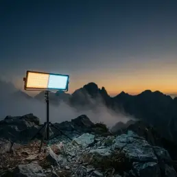 Bi-colour LED panel on a tripod glowing warm amber and cool daylight on a rocky mountain ridge at dusk, mist drifting across jagged peaks.