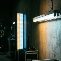 Bi-colour LED panel casting warm amber and cool daylight tones beside a fluorescent tube on a cinematic concrete studio wall.