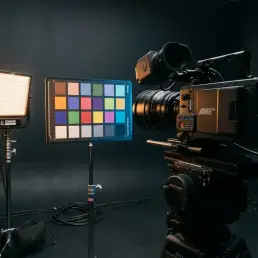 Cinema camera on tripod aimed at a color chart lit by a bi-colour LED panel casting warm amber to cool blue gradient across swatches in a dark studio.