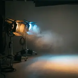 Two cinema lights on a studio floor casting contrasting warm amber and cool blue beams onto a gray backdrop, dust particles visible in the air.