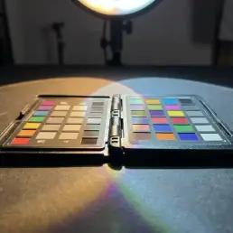 Color checker passport on studio surface lit by warm amber and cool daylight from a bi-colour LED, jewel-toned swatches casting crisp shadows.
