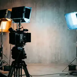 Cinema camera silhouetted before a concrete studio wall lit by overlapping warm amber and cool blue LED fixtures on a film set.