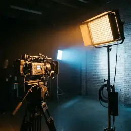 Professional LED panel splitting warm amber and cool daylight tones across a dark film set, illuminating a cinema camera on a tripod beside an exposed brick wall.