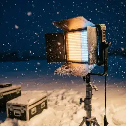 Bi-colour LED panel light on a stand casting warm amber and cool daylight tones over a snow-covered outdoor film set at dusk.