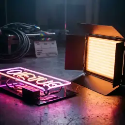 LED bi-colour panel light glowing amber beside a vintage neon sign on a dark film set, with cables and clapperboard in the background.