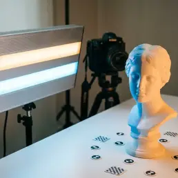 Bi-colour LED panel casting warm amber and cool blue light on a white ceramic sculpture during photogrammetry scanning, reference markers on studio surface.