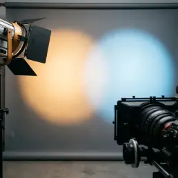 LED studio light projecting warm amber and cool daylight beams onto grey backdrop, cinema camera in foreground, dramatic color temperature contrast.