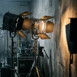 Two professional LED film lights on a studio set, one casting warm amber beam across textured concrete wall with dramatic shadows.