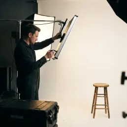 Fotógrafo ajustando un panel LED en un jirafa de estudio, con un fondo blanco y un taburete de madera vintage en primer plano.